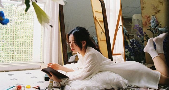 fair skinned Asian woman lying on her stomach and reading a book in front of a floor length mirror.jpg.optimal